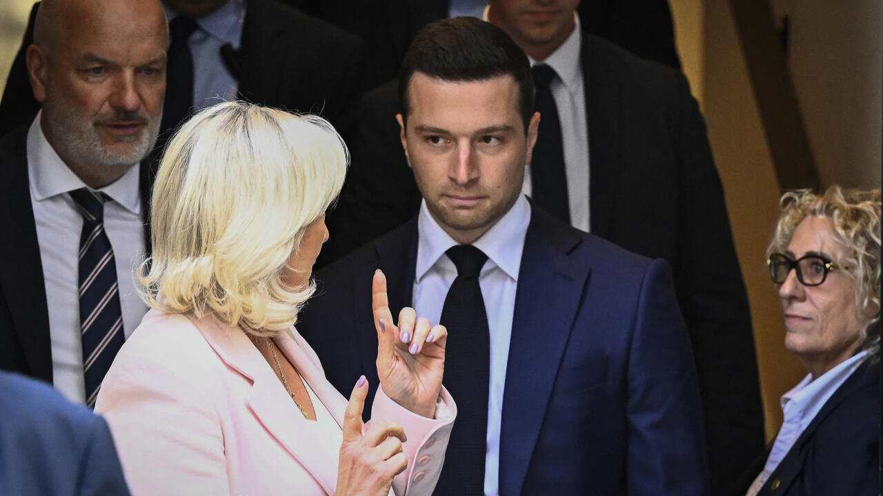 A blonde woman in a pale pink jacket gestures toward a man in a dark suit amid a group.