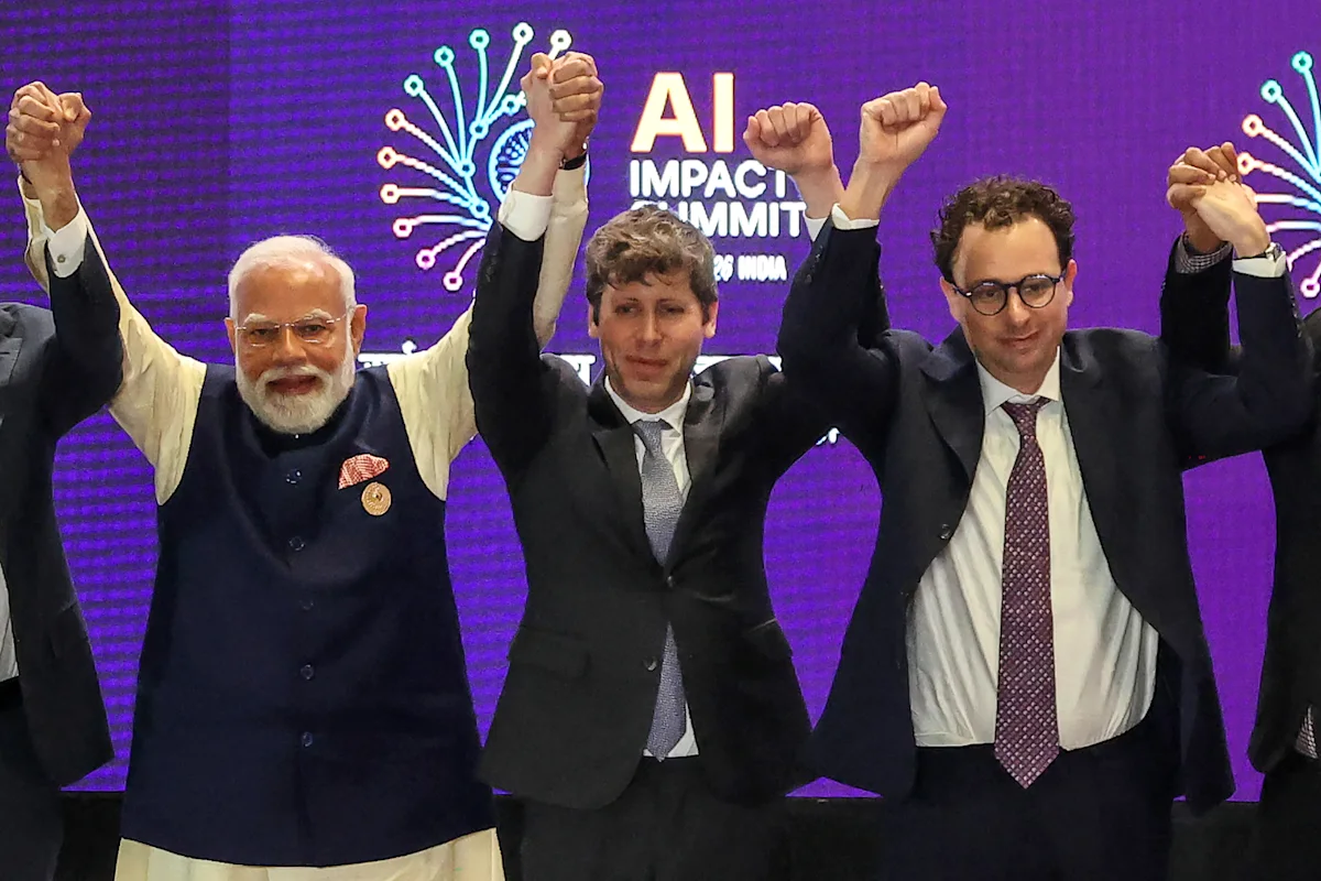 Three suited men raise joined hands onstage before a purple AI Impact Summit backdrop.