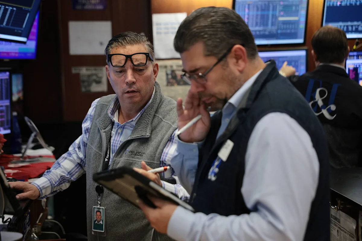 Stock market traders confer on a busy trading floor surrounded by financial monitors.