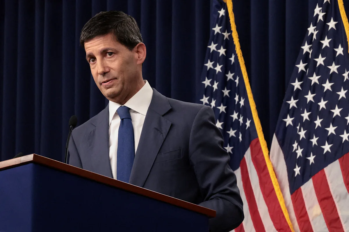 Man in suit speaking at a podium with American flags and dark curtains behind him