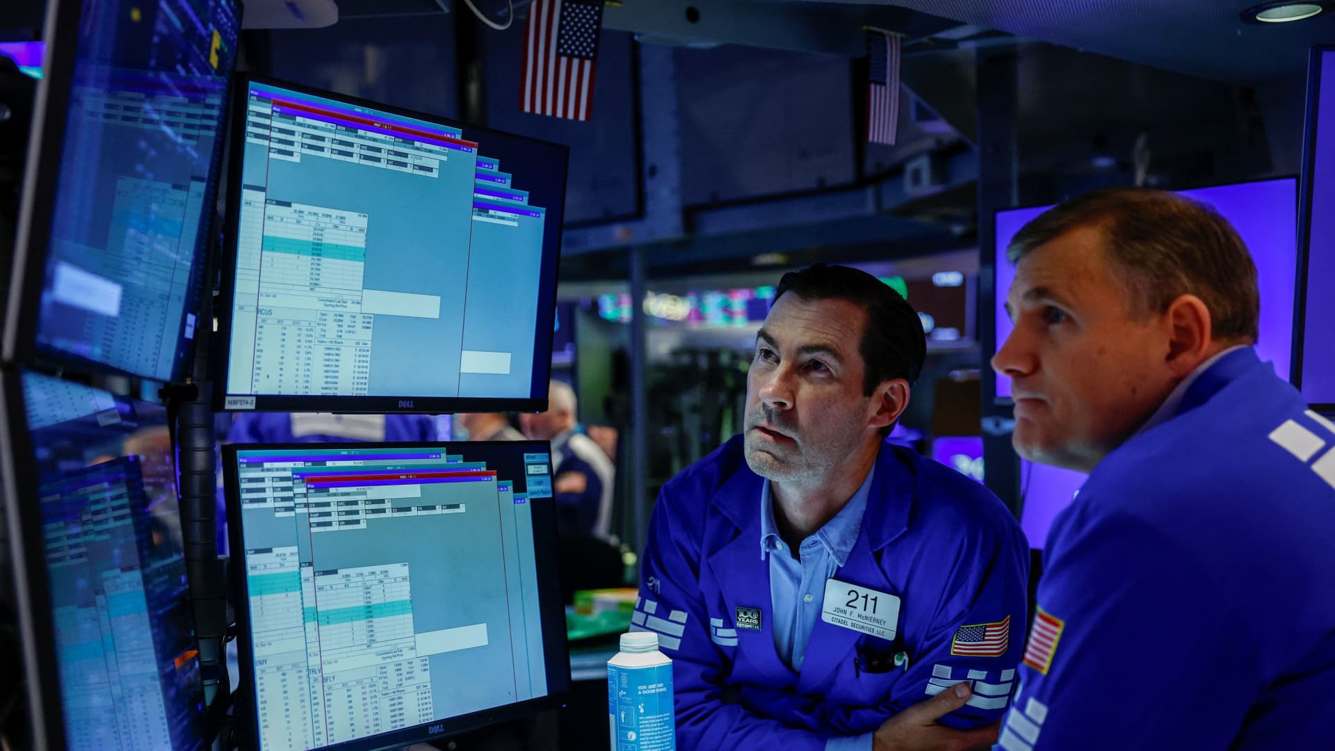 Two stock traders confer beside computer monitors displaying financial market data on a trading floor.