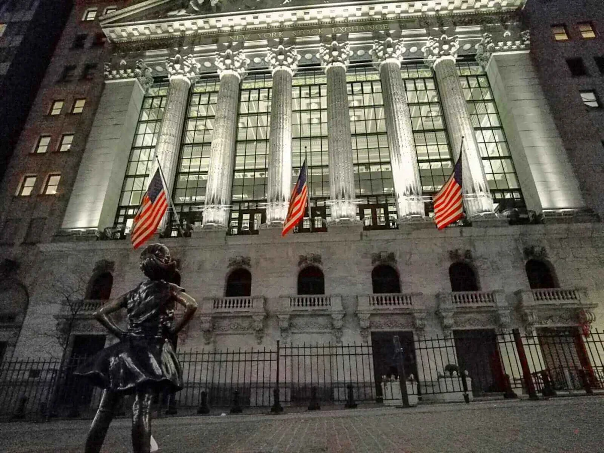 New York Stock Exchange facade with American flags and Fearless Girl statue at night.