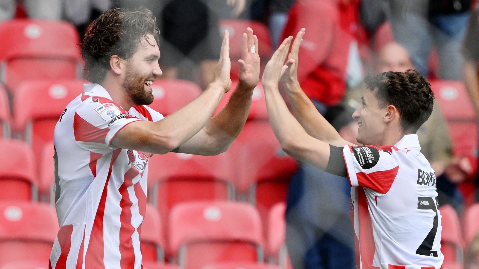 Two footballers in red-and-white shirts high-five in front of empty red stadium seats