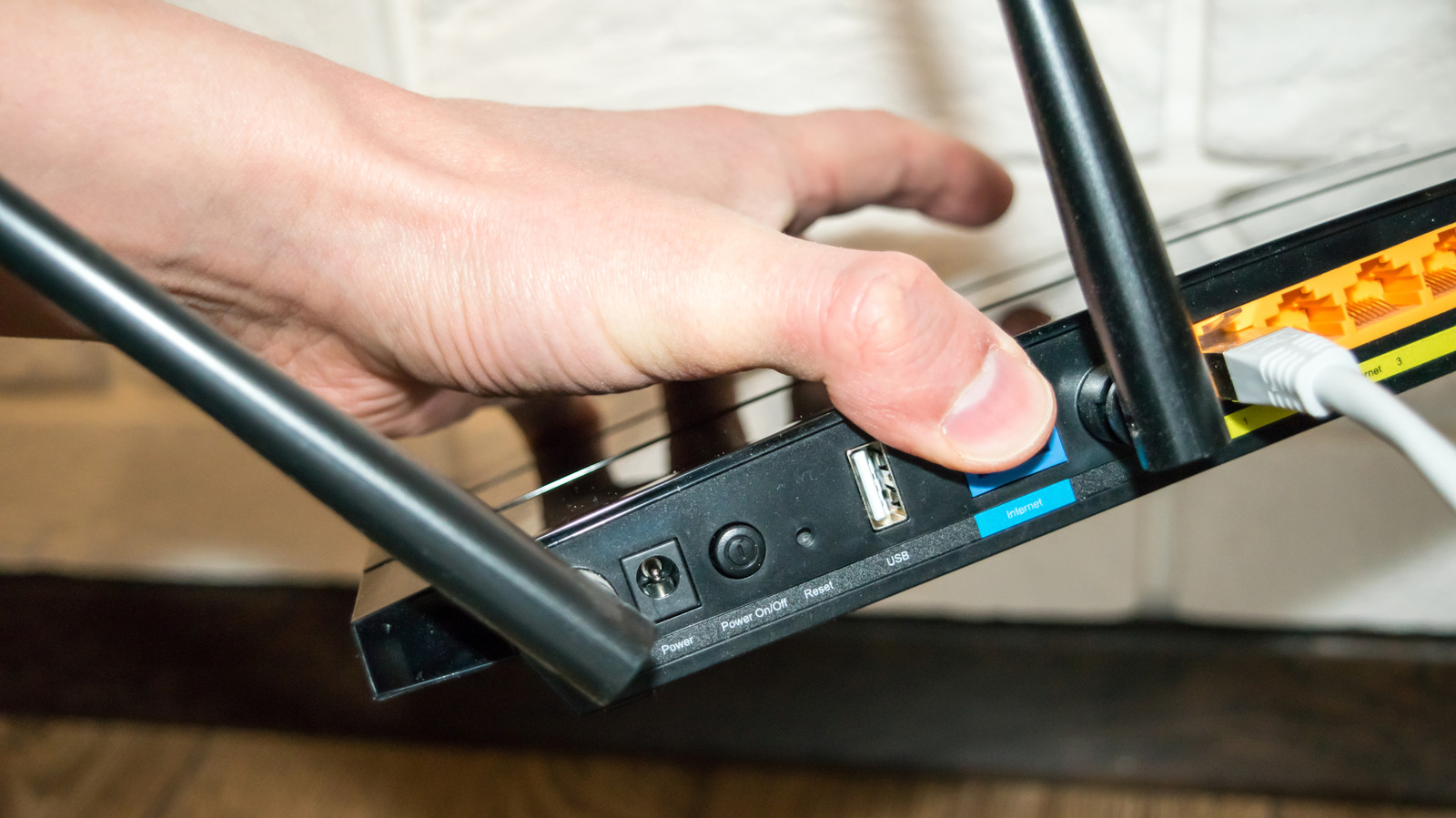 Hand pressing a button on a black Wi-Fi router beside its USB port and cables