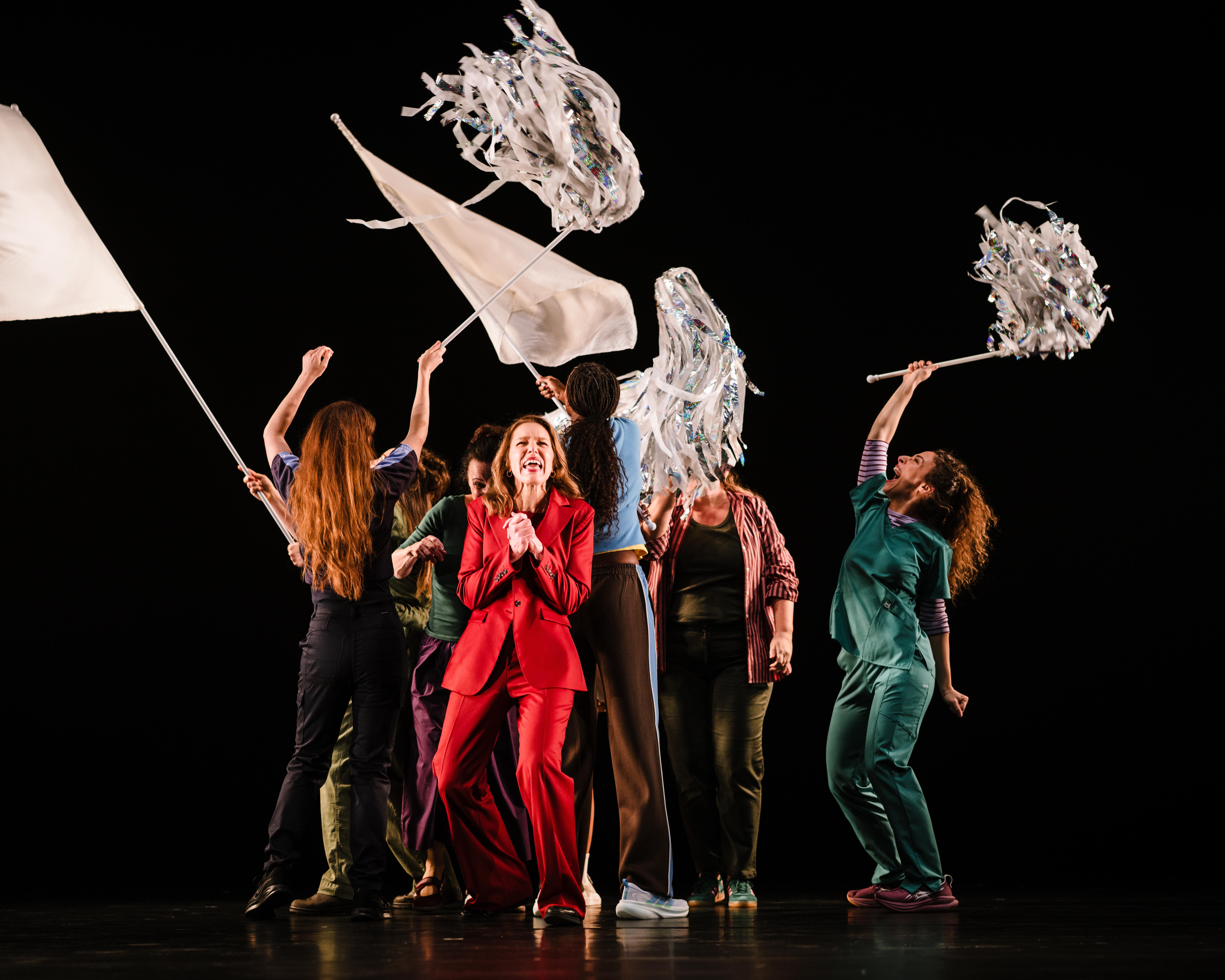Performers in colorful clothing raise white flags and shimmering streamers on a dark stage.