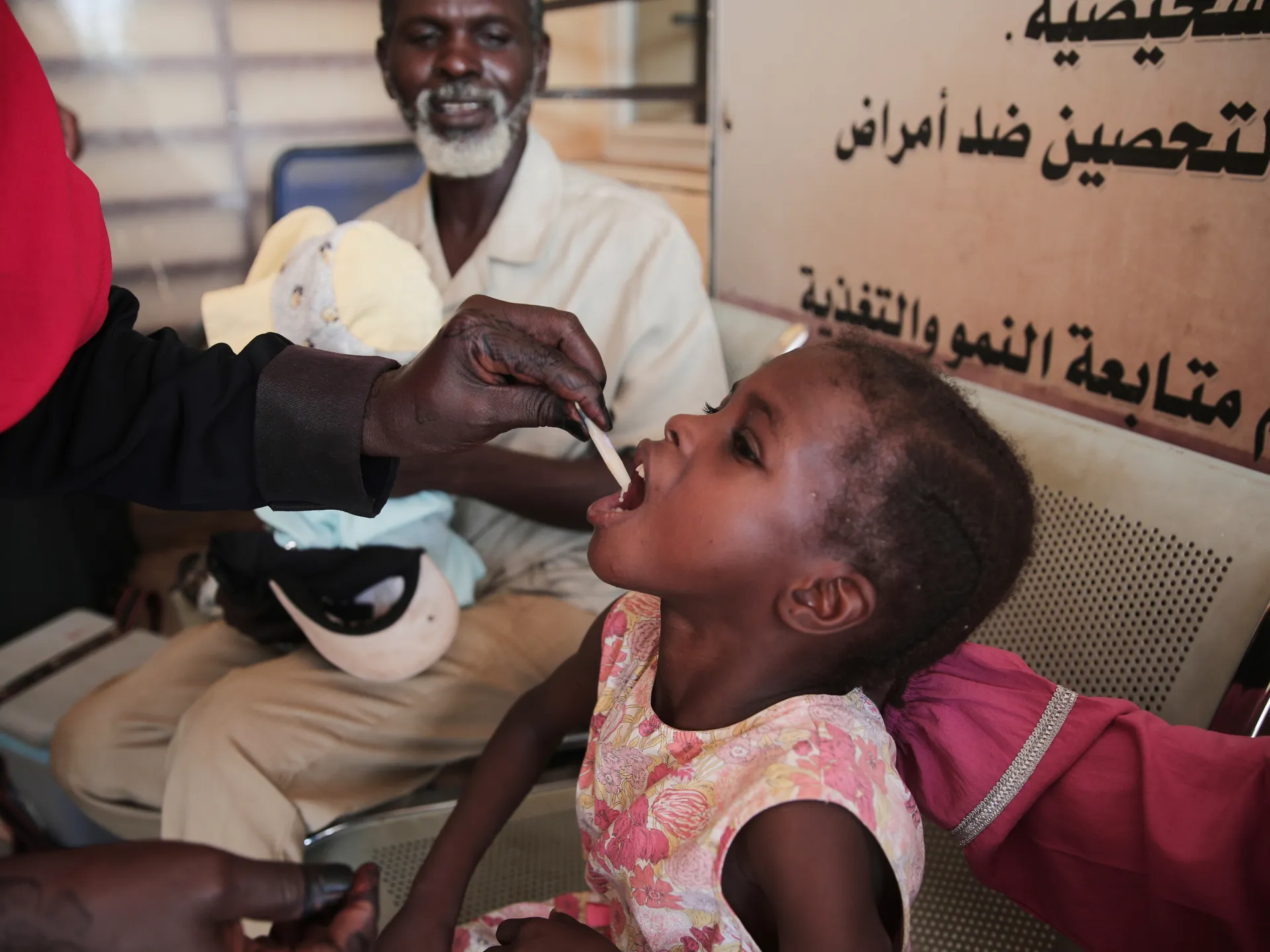 Child receives oral medicine from a health worker while adults sit nearby in a clinic.