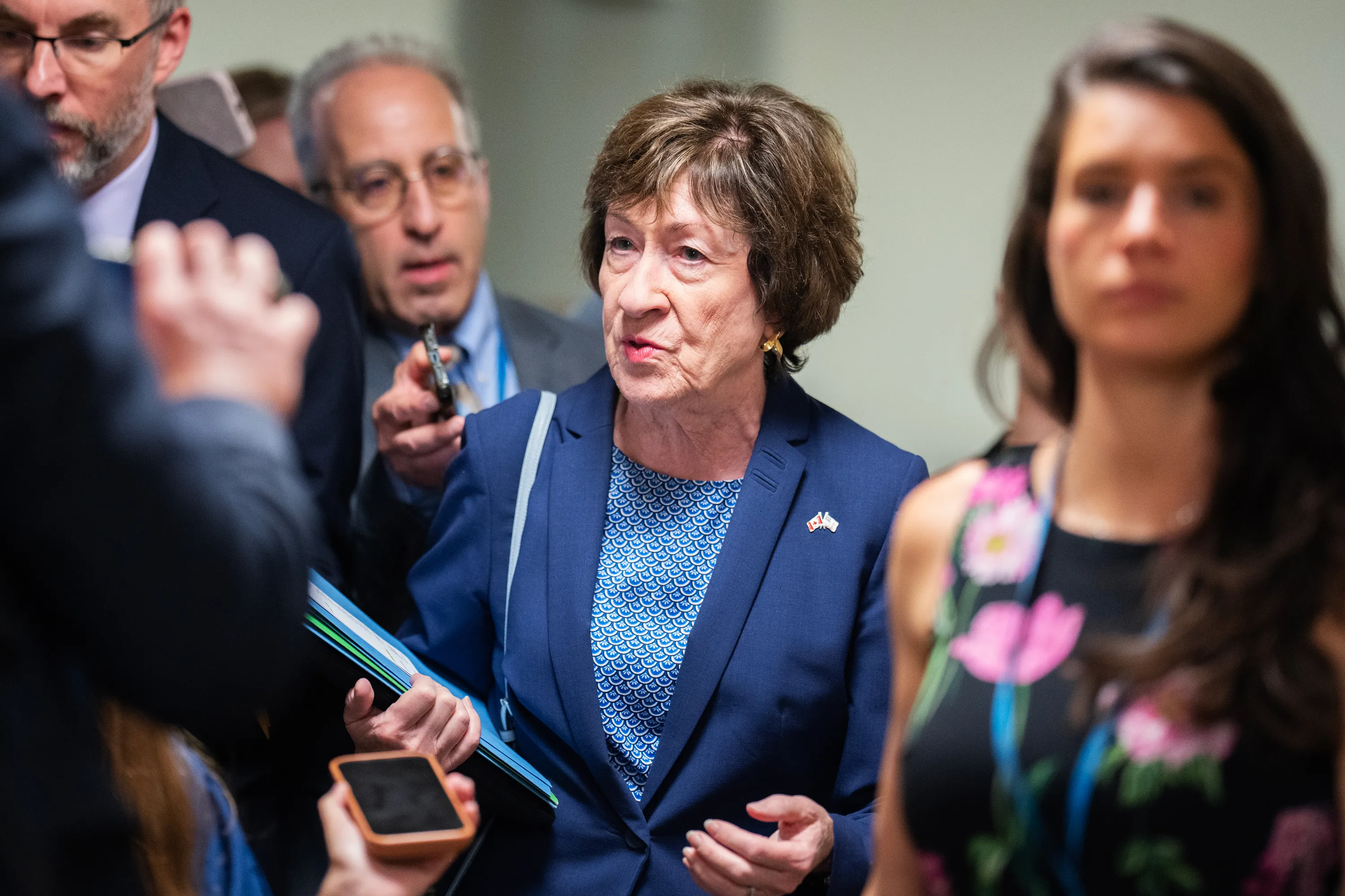 A woman in a blue suit stands among reporters, with microphones and phones in the foreground.