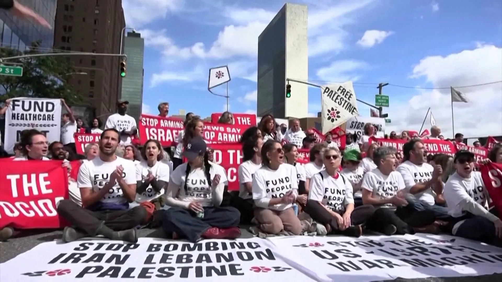 A seated protest crowd holds red and white signs, with a banner spread across the foreground.