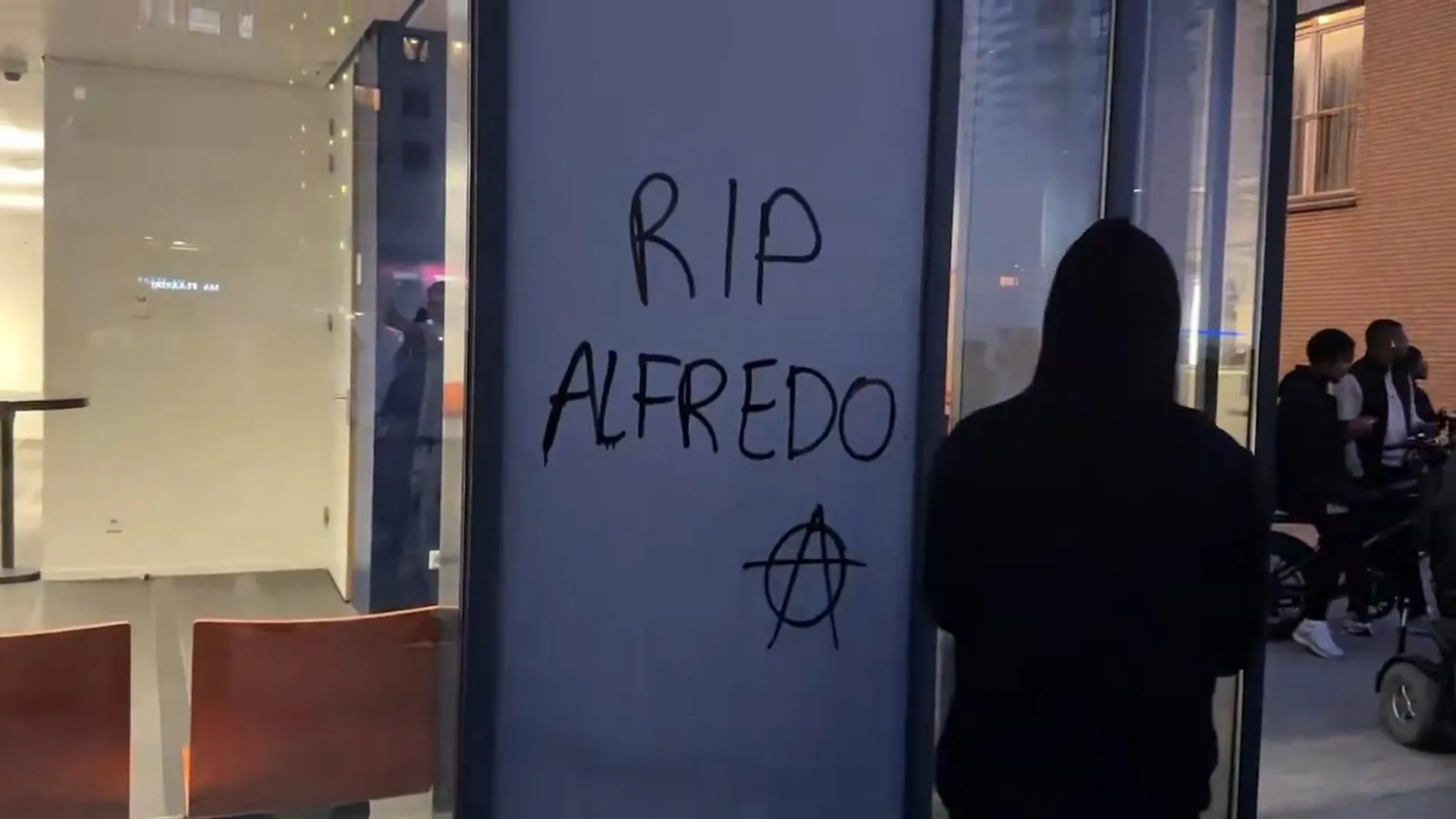 Graffiti reading “RIP Alfredo” on a building entrance as people gather outside.