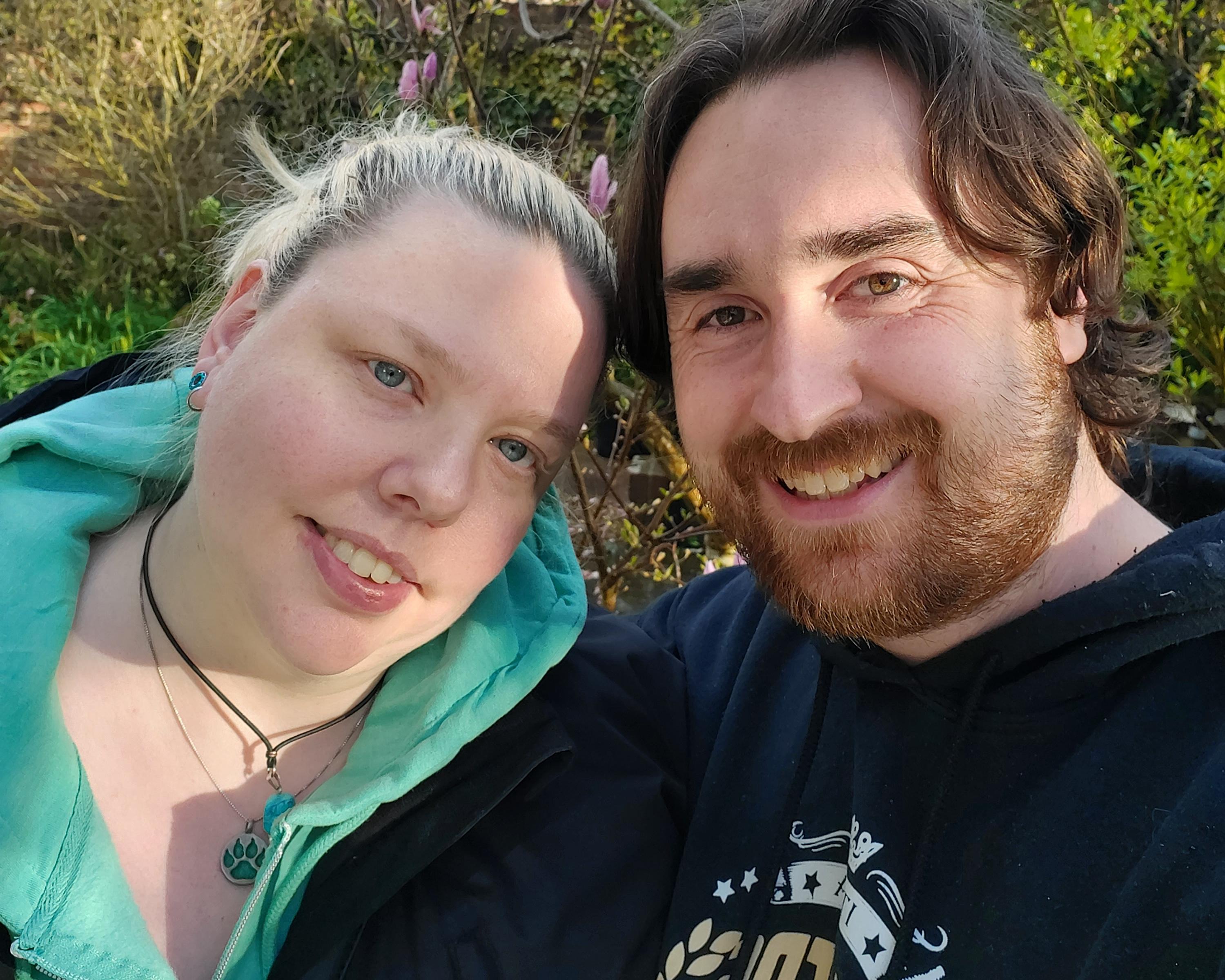 Smiling couple posing for a close-up selfie outdoors amid green foliage and flowering plants.