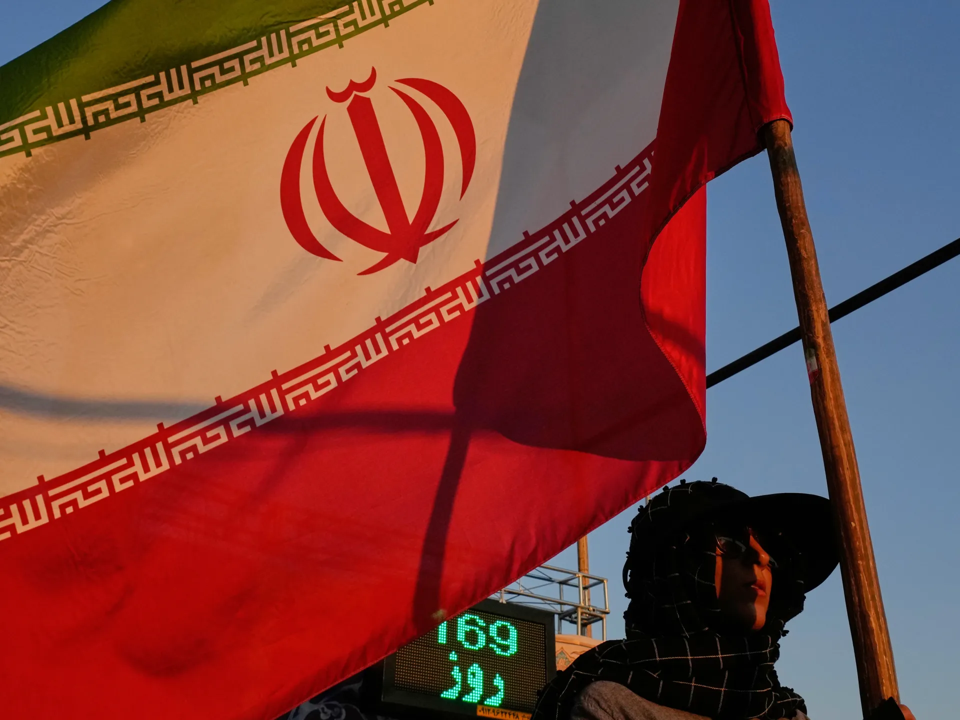 Iranian flag billows beside a person wearing dark clothing and a head covering outdoors.