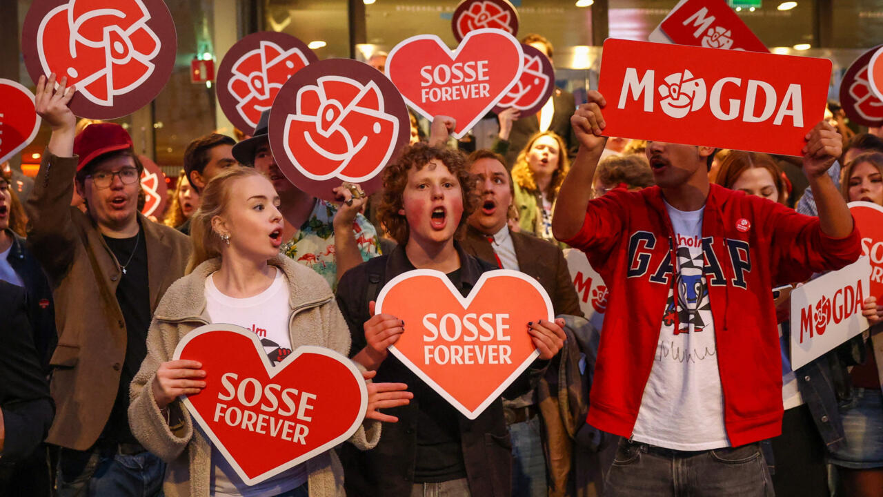Crowd holds red hearts and political signs, including “SOSSE FOREVER” and “M GDA,” during a rally.