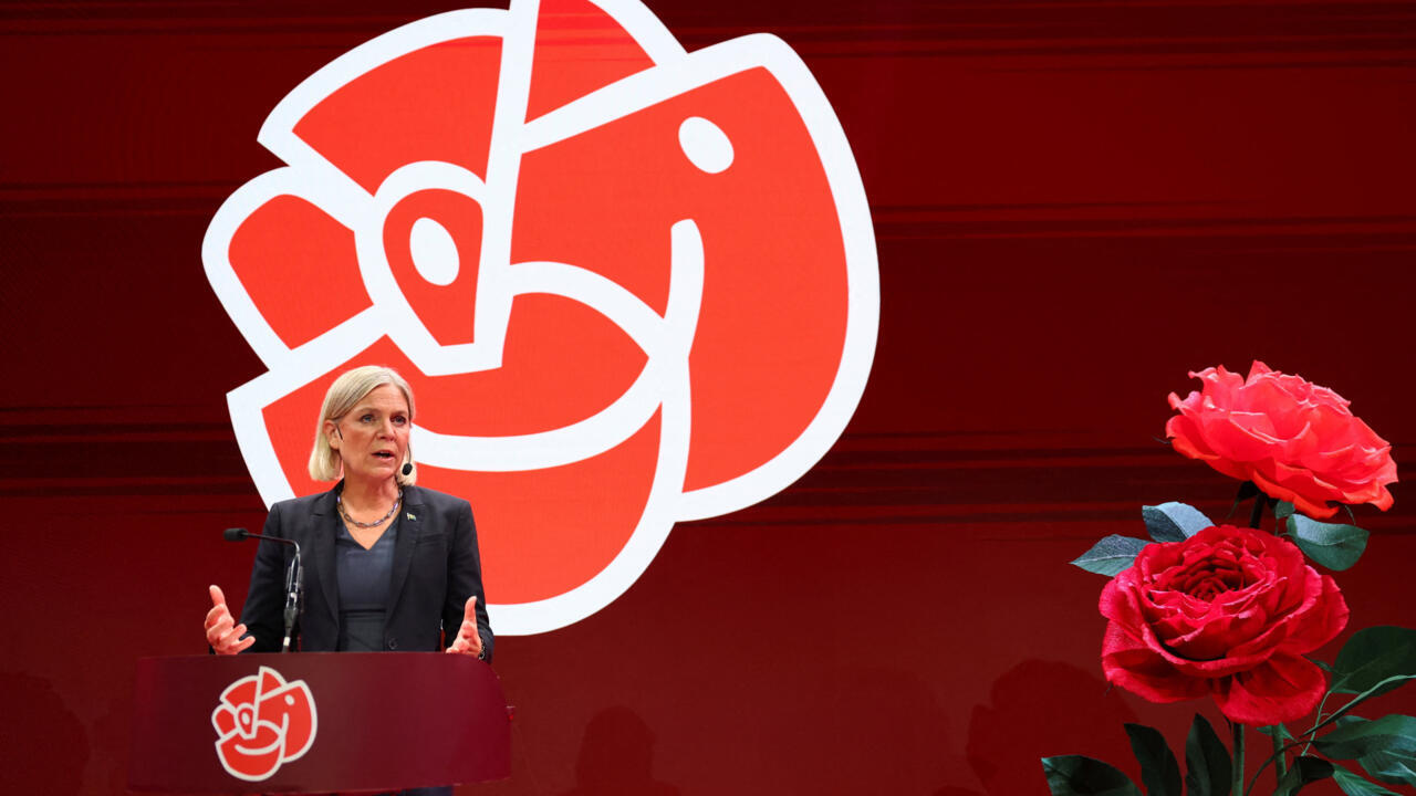 Blonde woman speaks at a podium before a large red political party logo, with roses beside her.