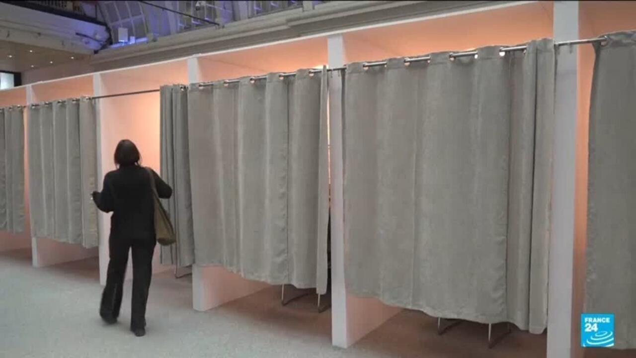 A voter walks past curtained polling booths in an indoor voting venue.