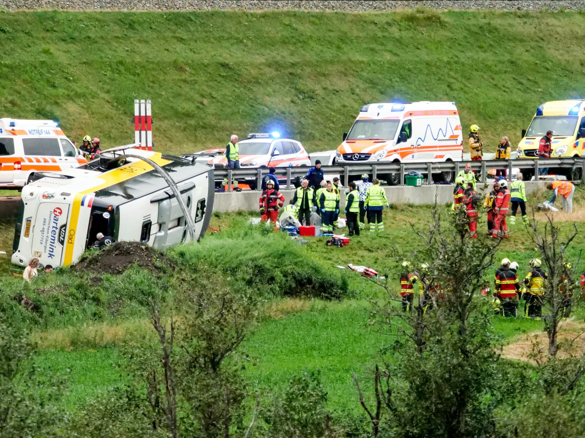 Emergency crews attend an overturned tour bus beside ambulances on a grassy roadside.
