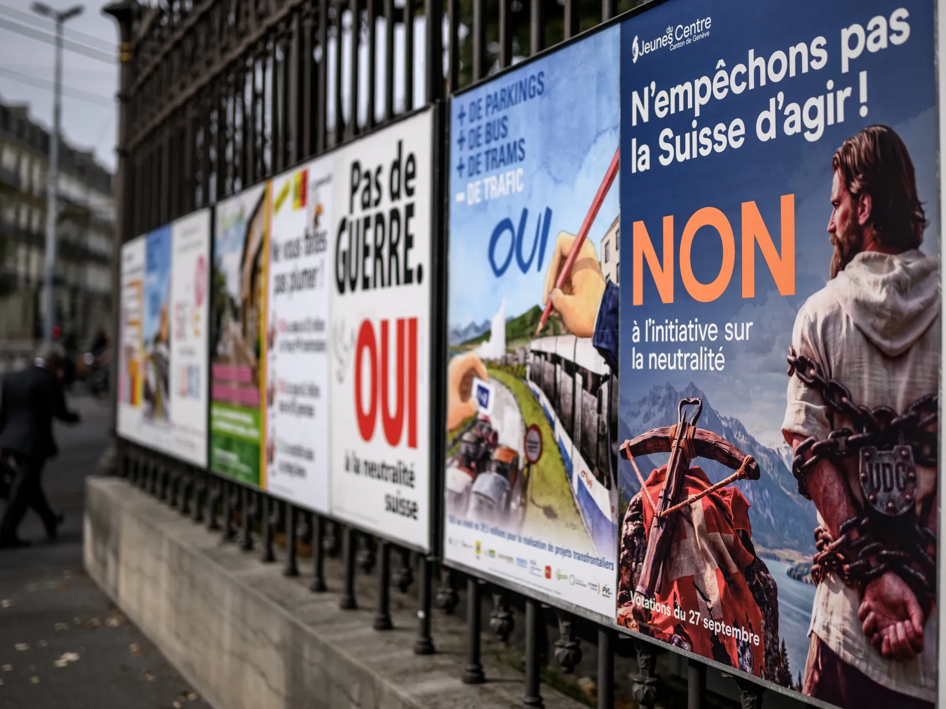 Swiss referendum posters line a sidewalk, including a blue neutrality poster with large orange “NON” lettering.
