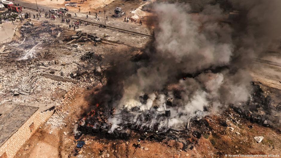 Aerial view of smoldering wreckage, burning debris and thick smoke near a roadway.