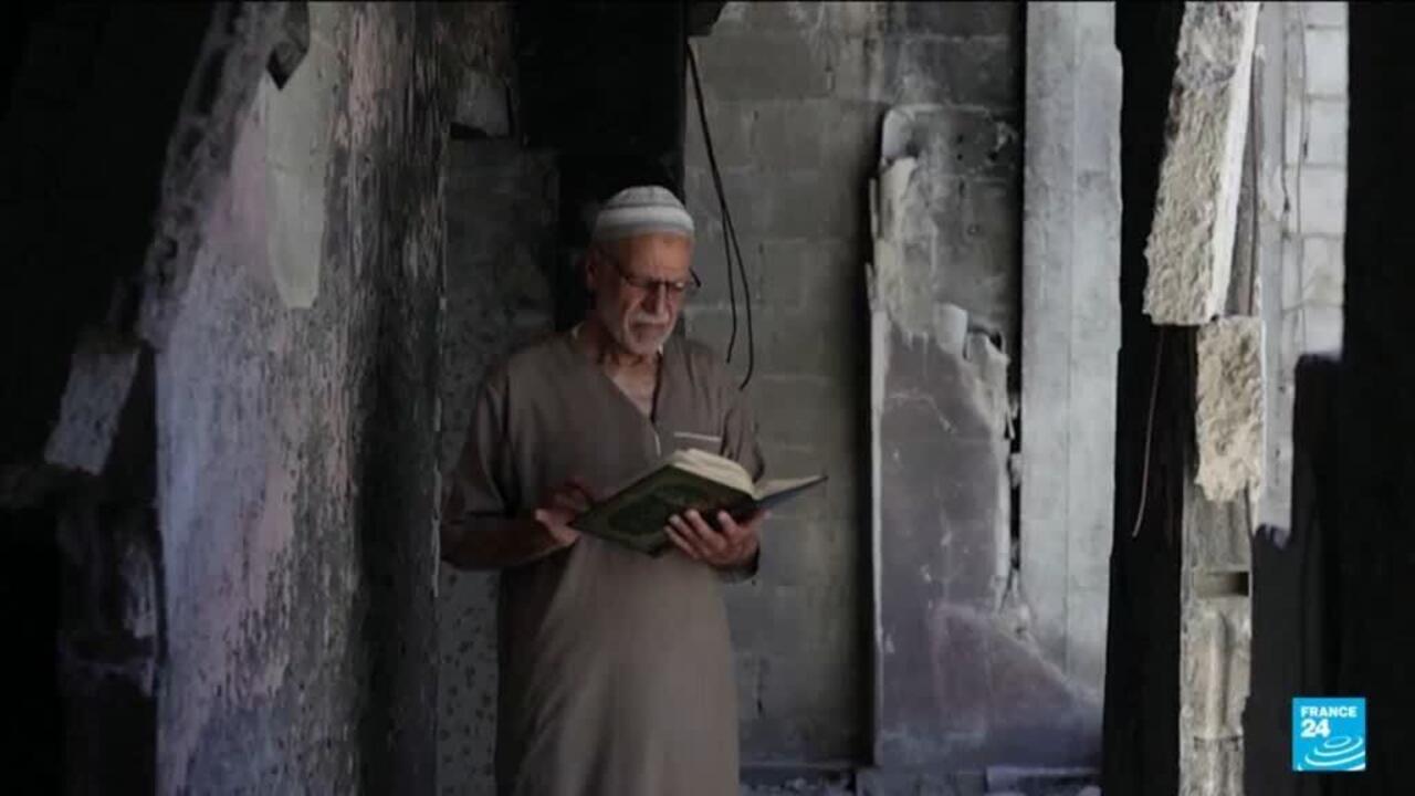 Older man in traditional cap reads an open book inside a damaged building.