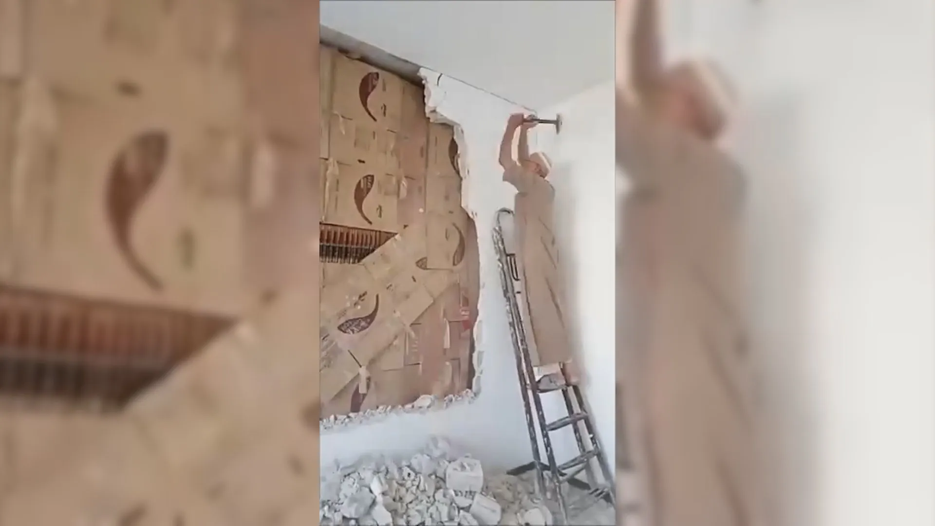 Man on ladder removes plaster from wall, revealing a concealed room filled with stacked cardboard boxes.