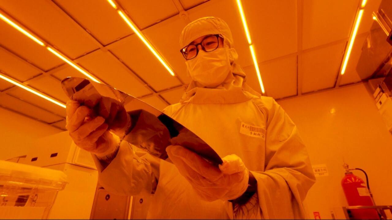 A masked worker in protective clothing holds a semiconductor wafer under bright orange lighting.