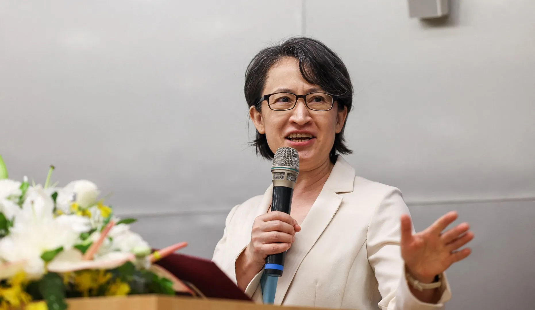 Woman in glasses and white blazer speaking into a microphone while gesturing at an event