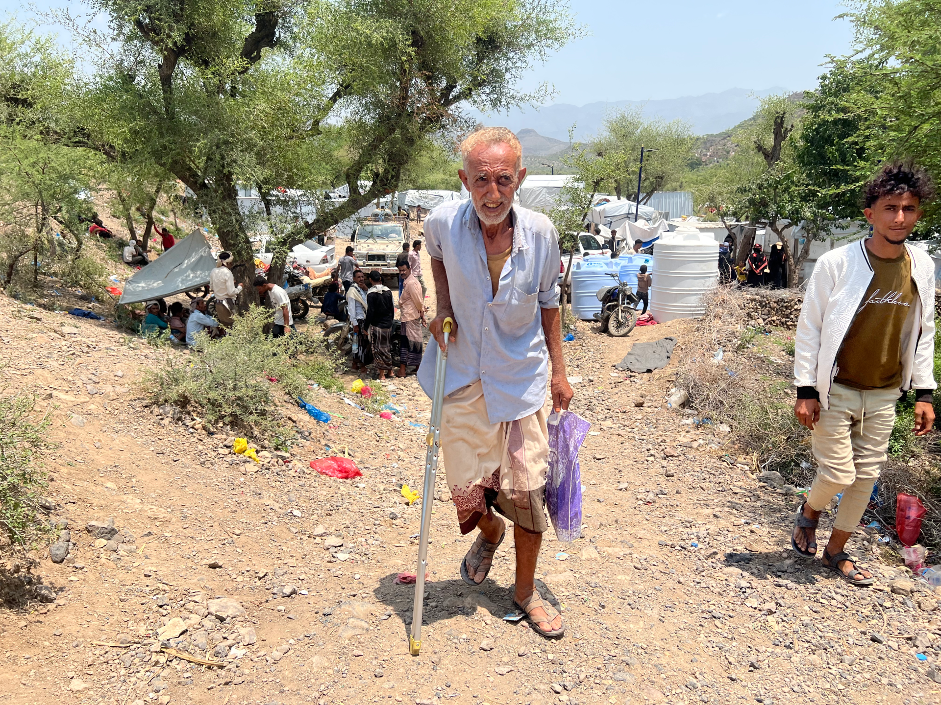 Older man using crutches walks through a crowded hillside camp with tents and water tanks.
