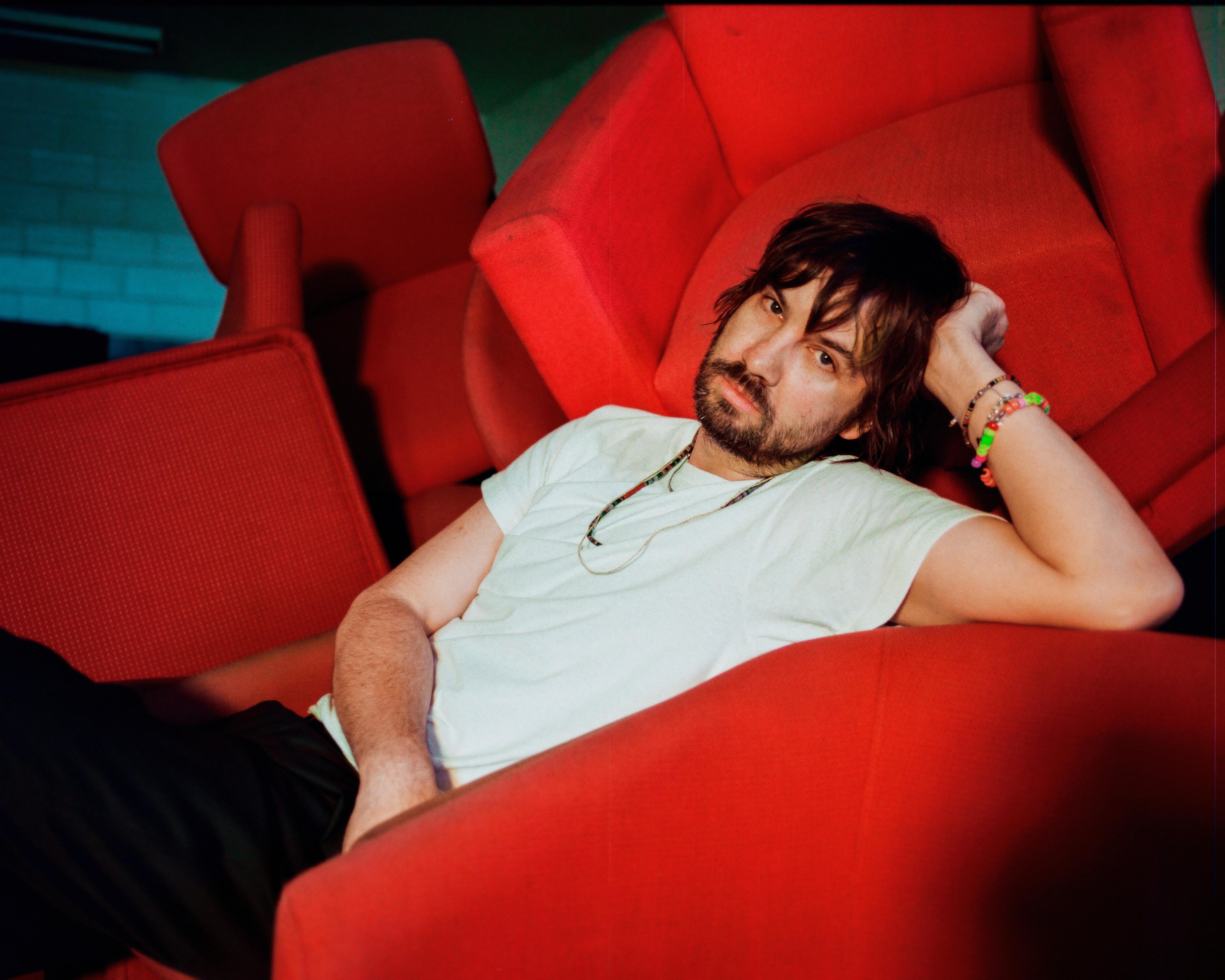 Bearded man in white shirt reclines across vivid red theater seats, looking toward the camera.