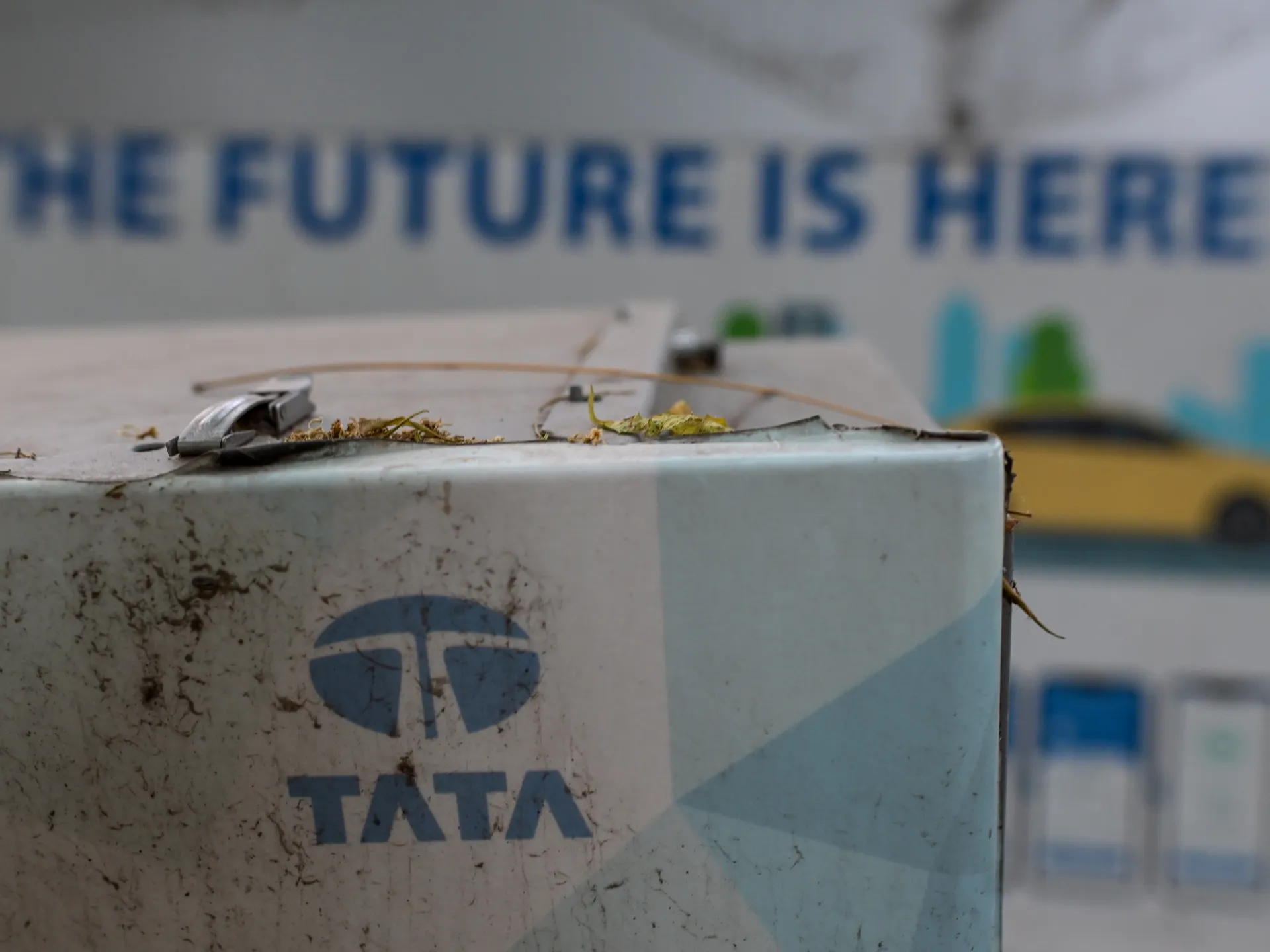 A blue Tata logo appears on a weathered wall beneath a blurred sign reading “THE FUTURE IS HERE.”