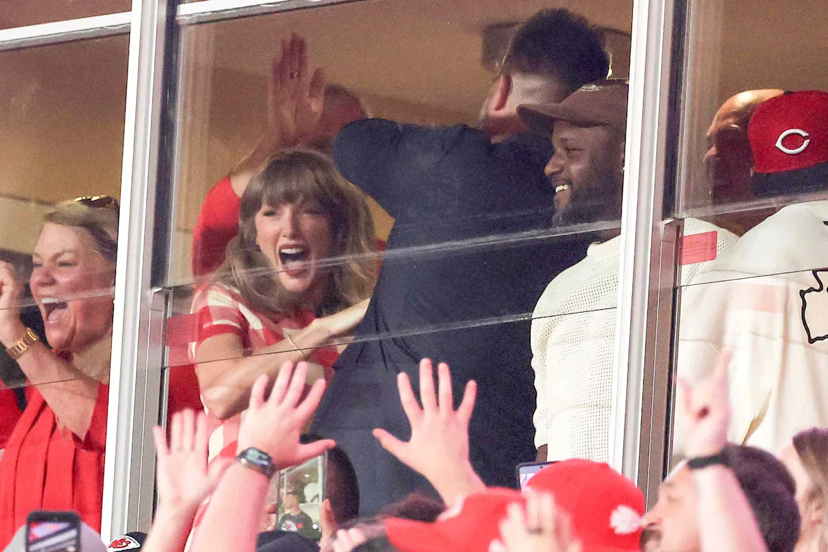 Taylor Swift celebrates in a stadium suite beside Jason Kelce as fans reach toward the glass.