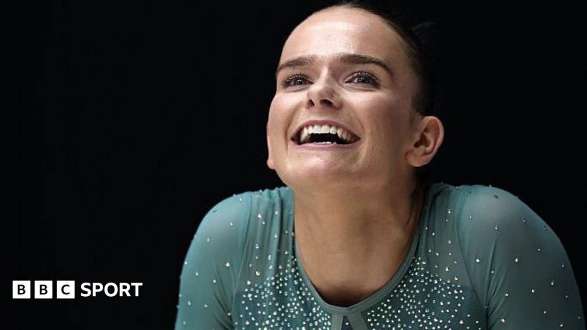 Smiling gymnast wearing a teal, crystal-studded costume against a dark background