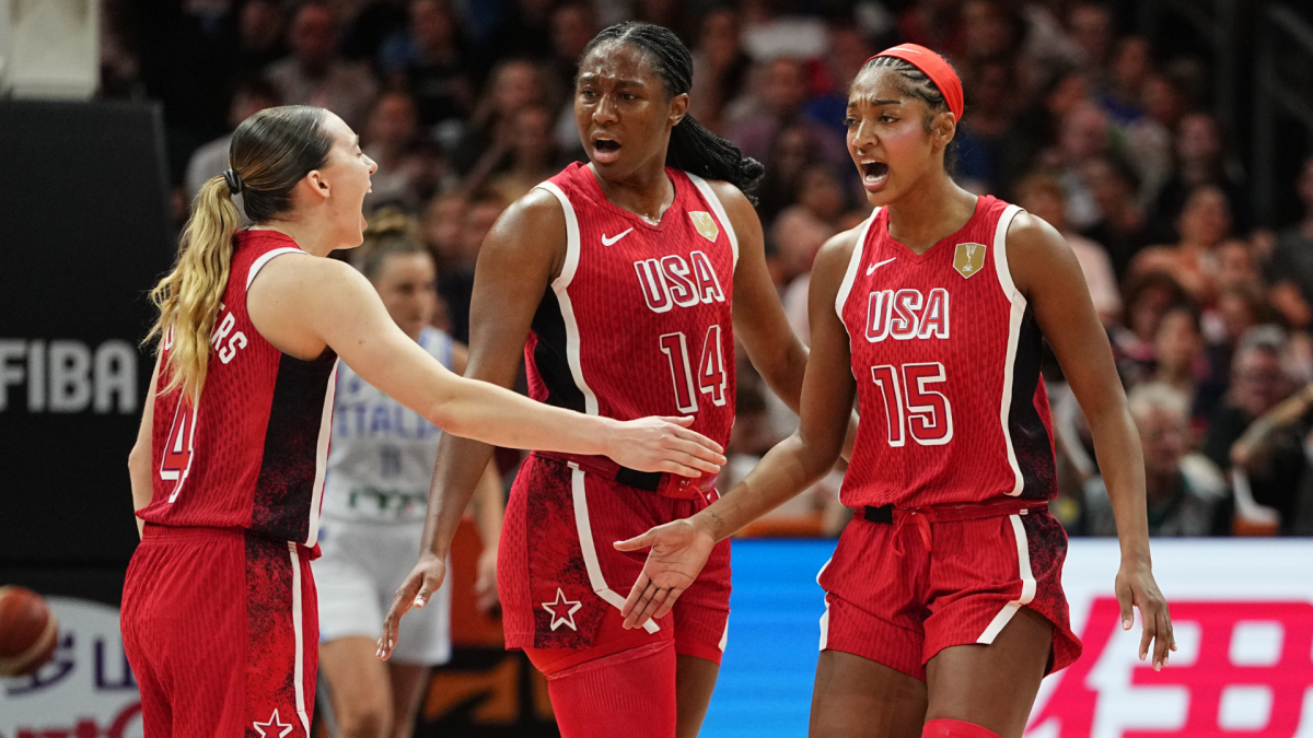 Three USA women’s basketball players in red uniforms gesture and talk during a game.