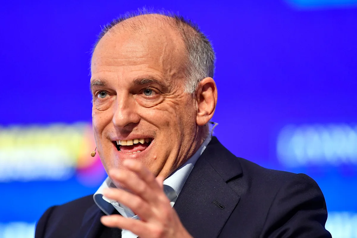 Bald man in dark suit speaking into a headset microphone against a blue background