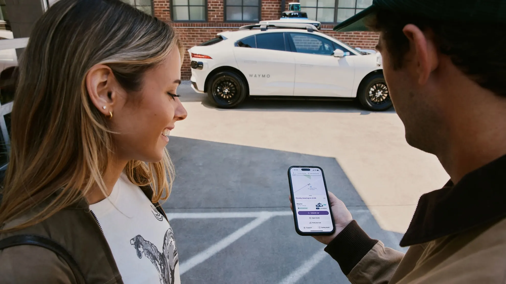 Two people view a smartphone beside a white Waymo autonomous vehicle.