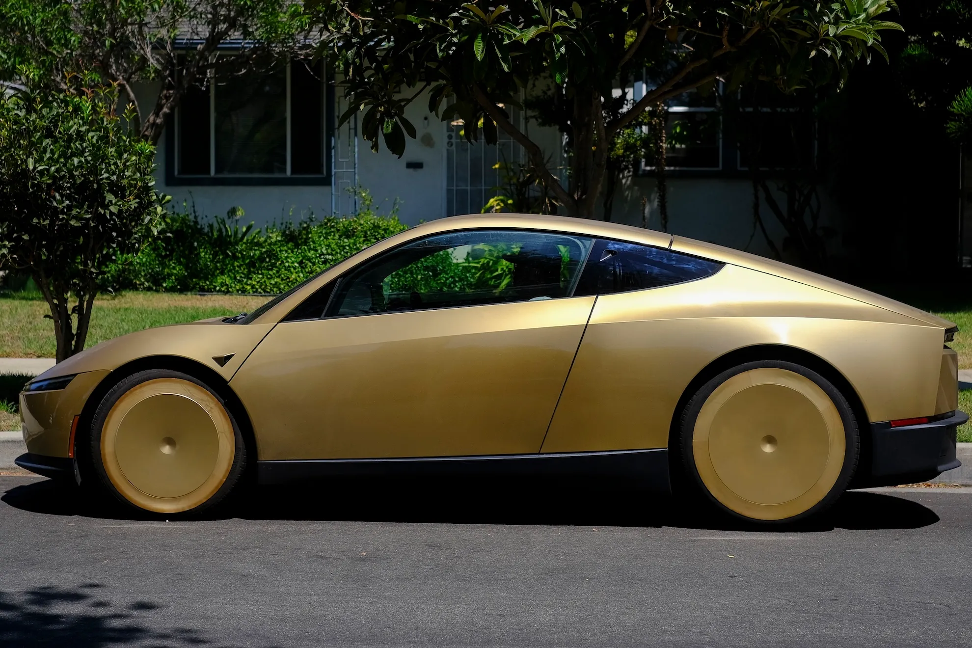 Gold futuristic Tesla Cybercab parked curbside in front of a leafy residential house.