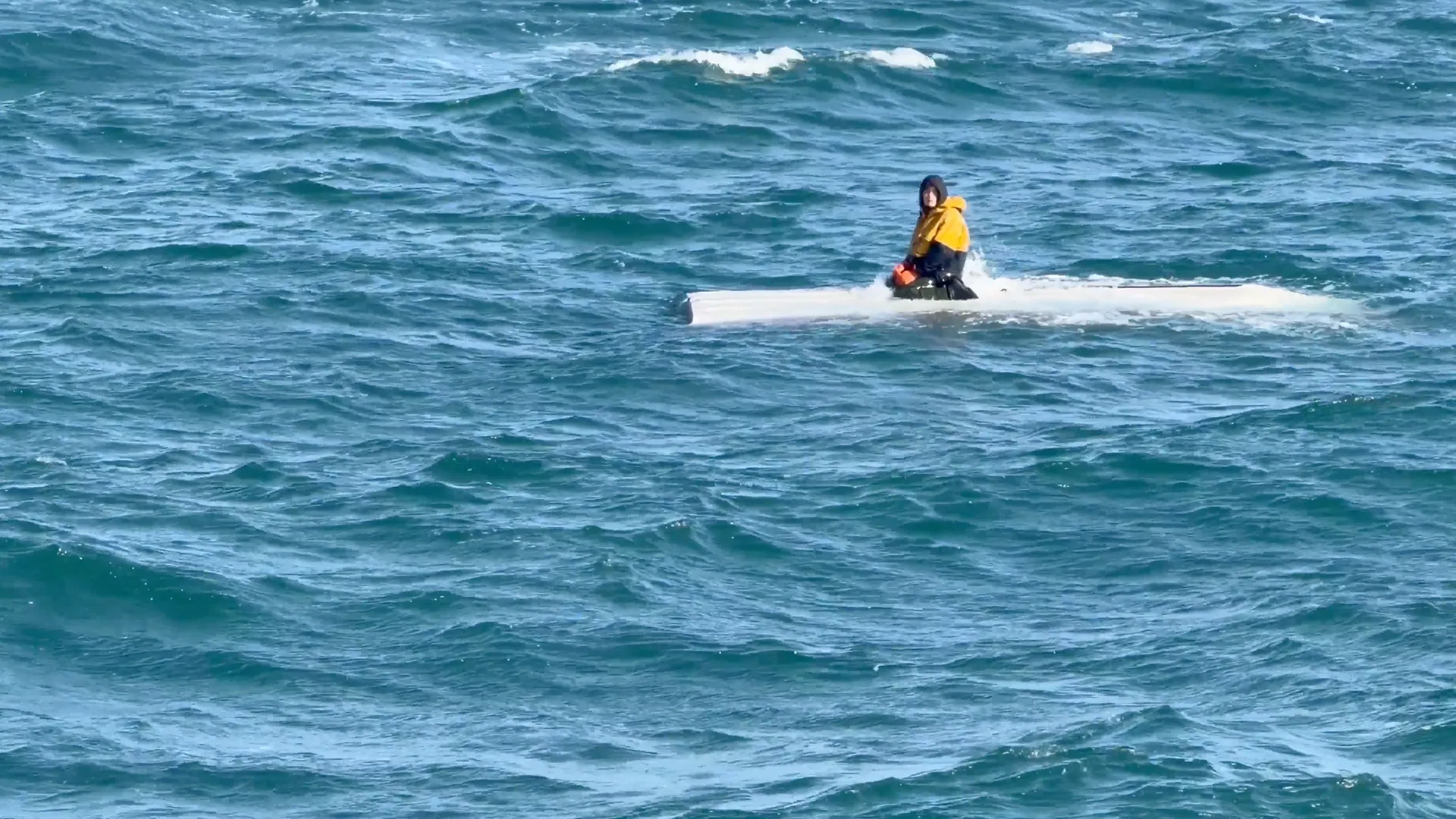 Person in yellow jacket sits on a white boat amid rough blue ocean waves.
