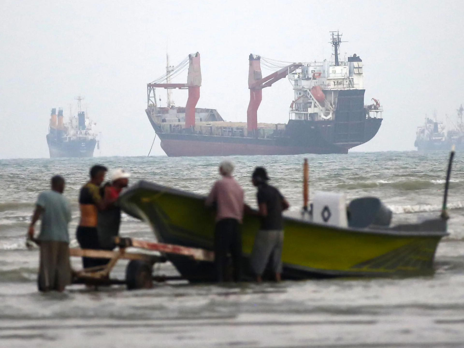 Workers push a small fishing boat through shallow surf with cargo ships offshore.
