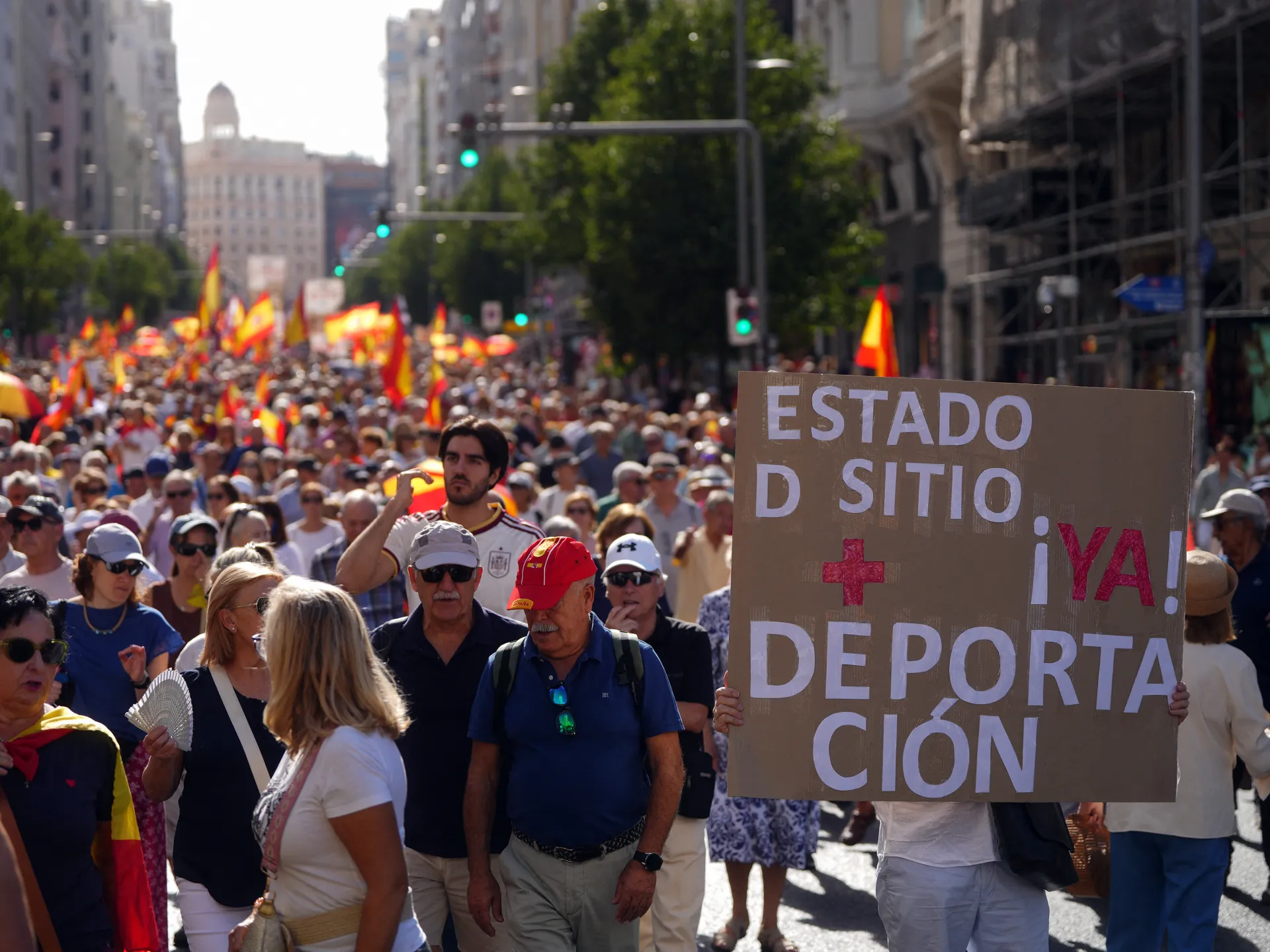 Crowd marches beneath Spanish flags, with a foreground protester holding a large cardboard sign.
