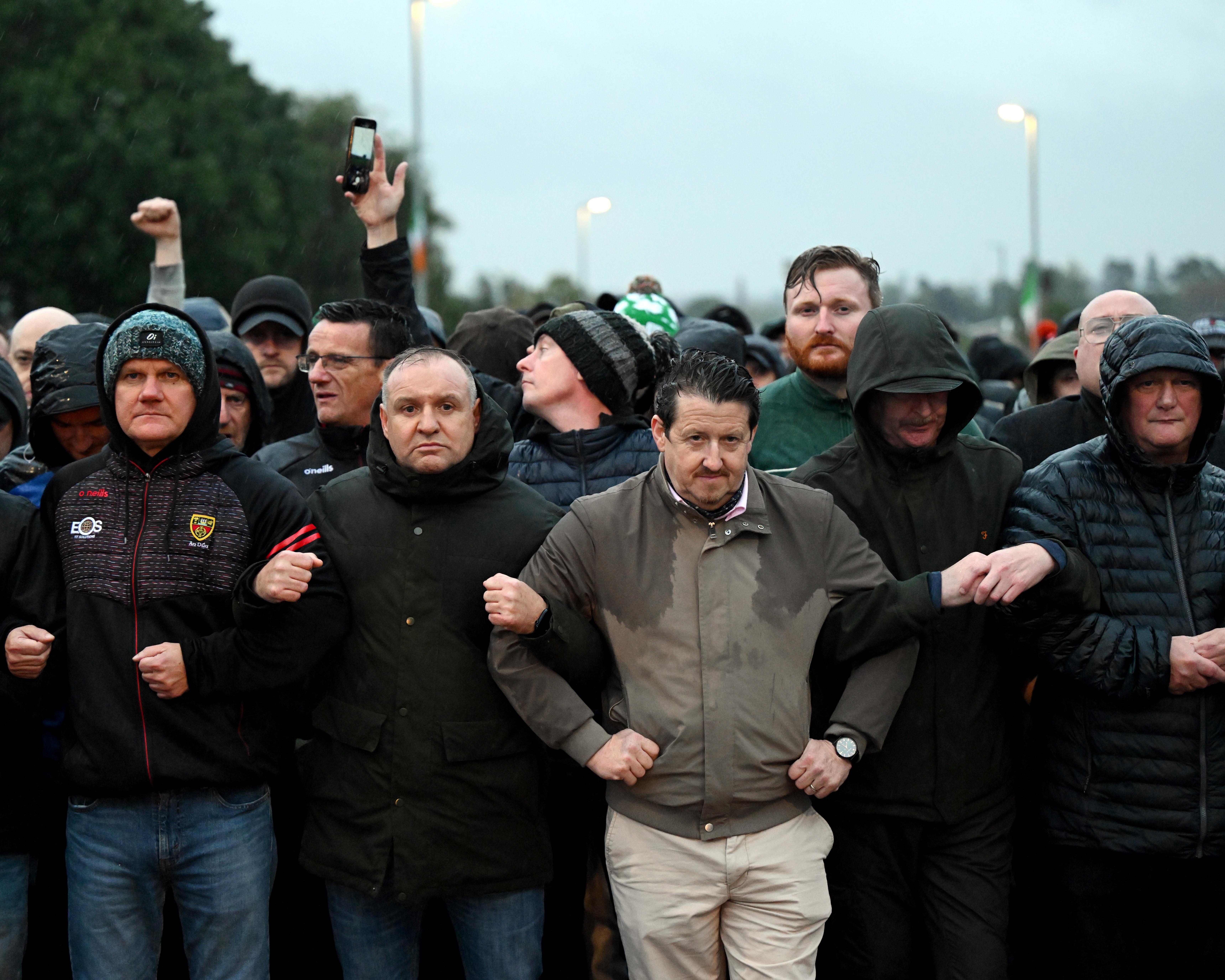 A group of men in rain jackets stand tightly packed, with one person holding a phone aloft.
