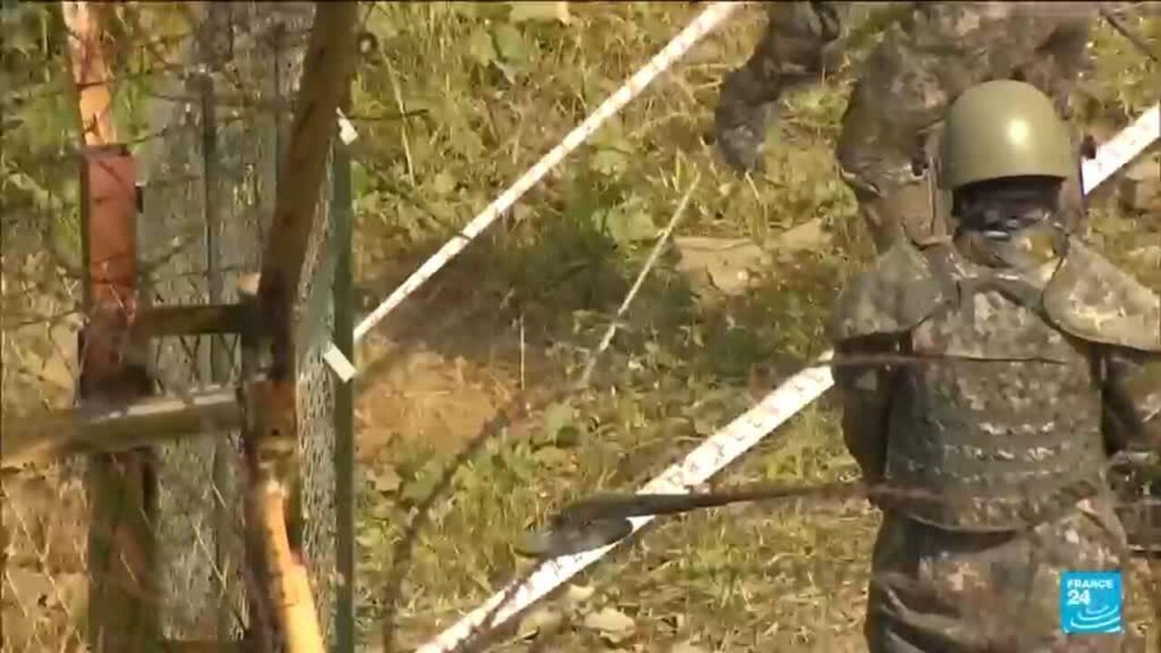 A helmeted soldier in camouflage stands beside a damaged fence on a wooded slope.