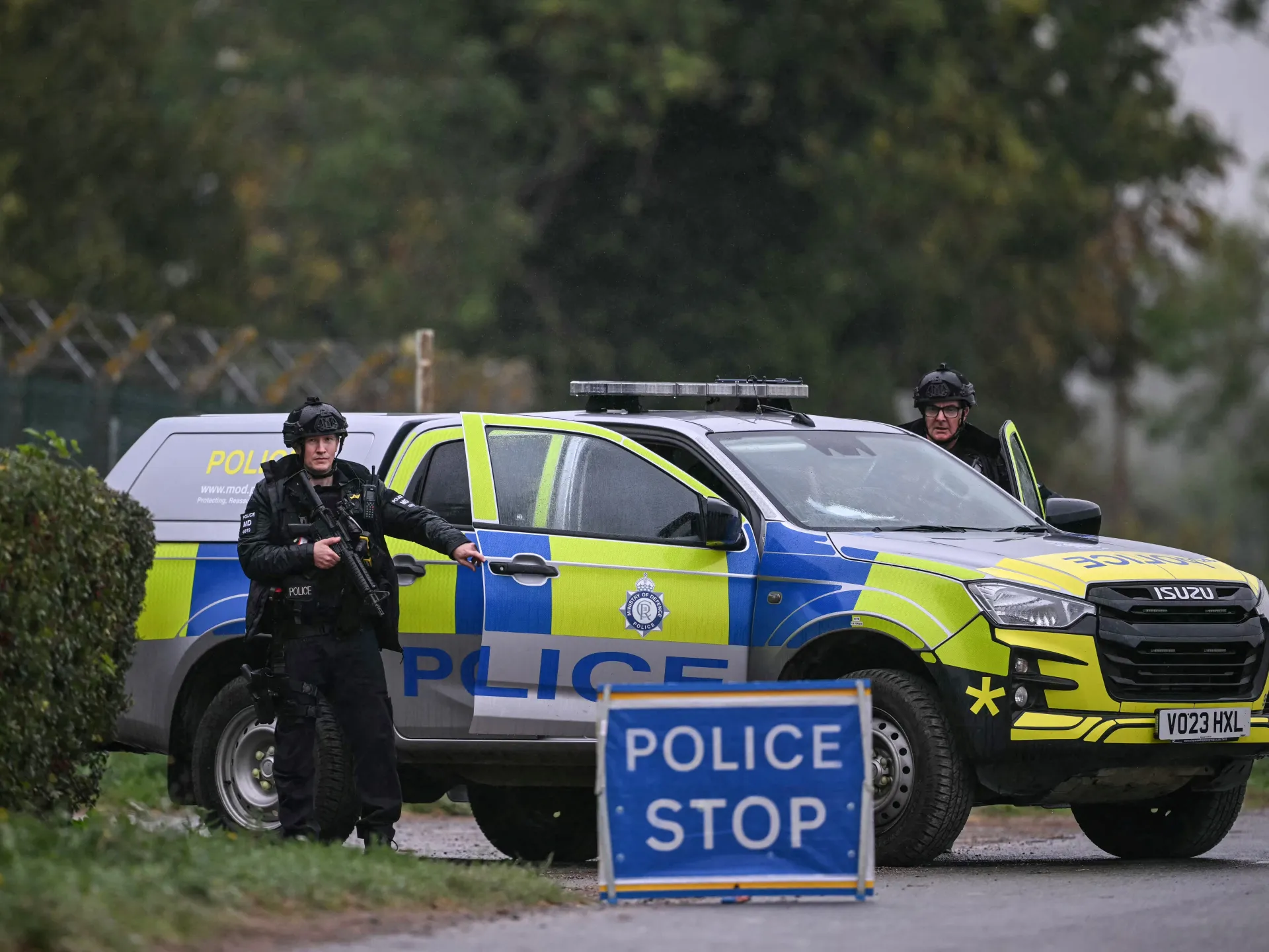 Armed officers stand beside a marked police vehicle and a police stop sign on a road.