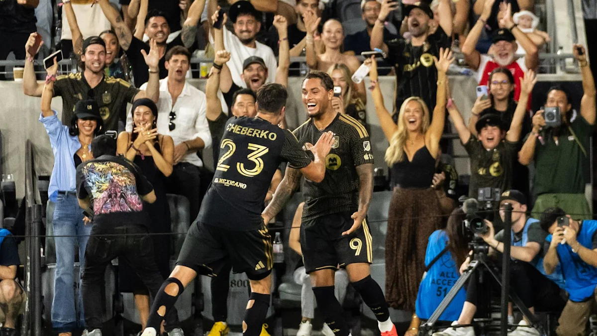 Two soccer players in black-and-gold kits celebrate in front of cheering supporters.