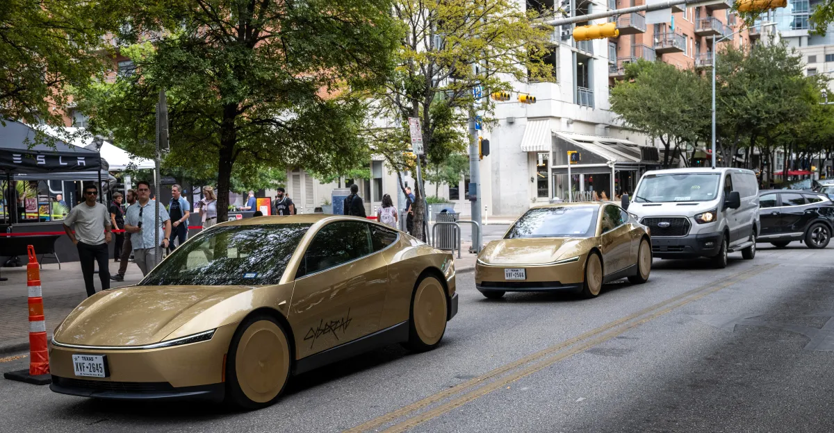 Two gold Tesla Cybercab vehicles travel along a city street near pedestrians and storefronts.