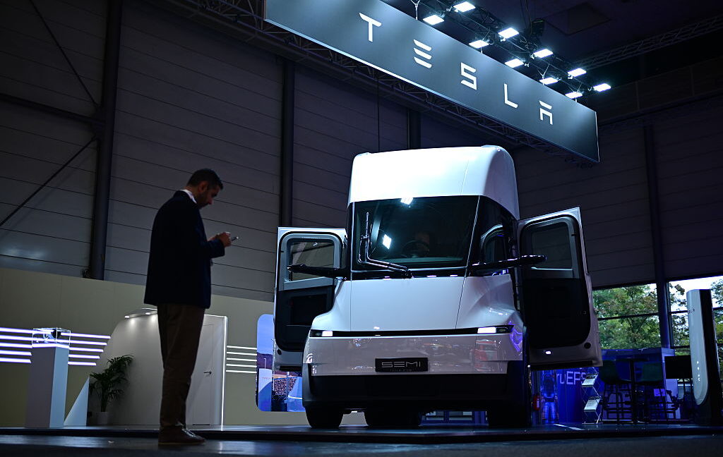 White Tesla Semi displayed indoors with both doors open as a man looks at a phone nearby.