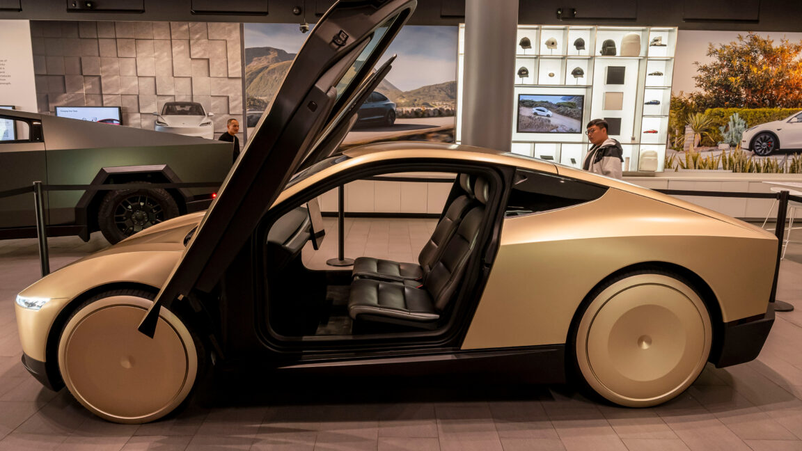 Futuristic gold vehicle displayed indoors with its upward-opening driver-side door raised.