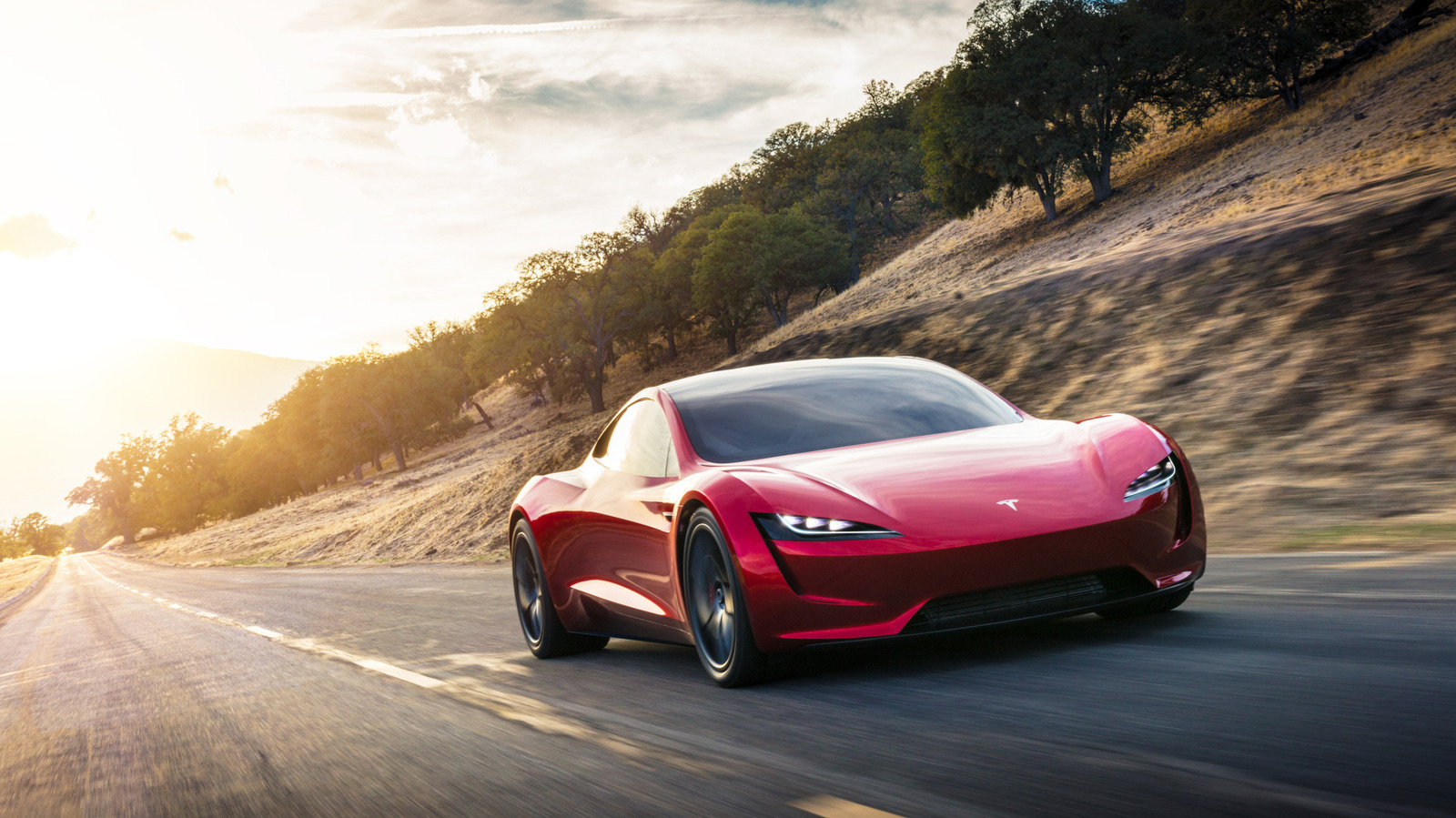 Red Tesla sports car driving along a sunlit winding road beside a wooded hillside