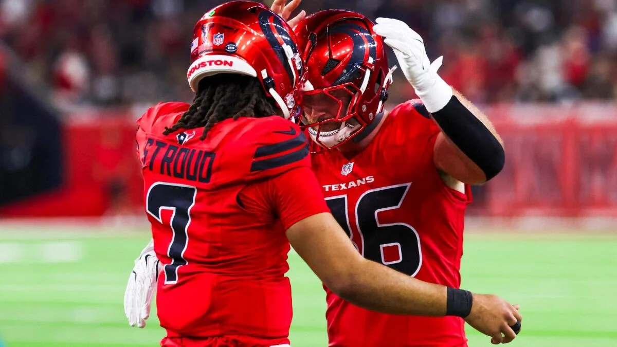 Two Houston Texans players in red uniforms celebrate together on the field, one raising a gloved hand.