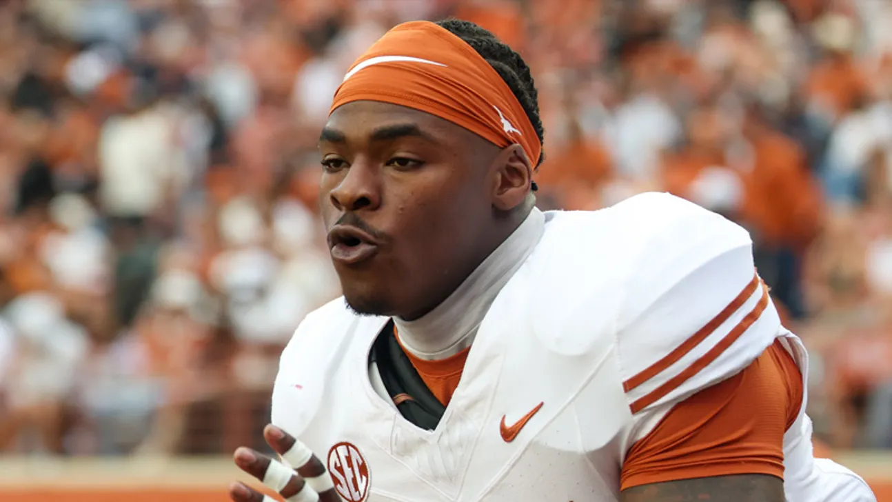 Football player in a white and orange uniform gestures in front of a blurred crowd.