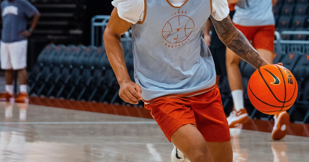 Basketball player in a white Texas practice shirt and orange shorts dribbles on court.