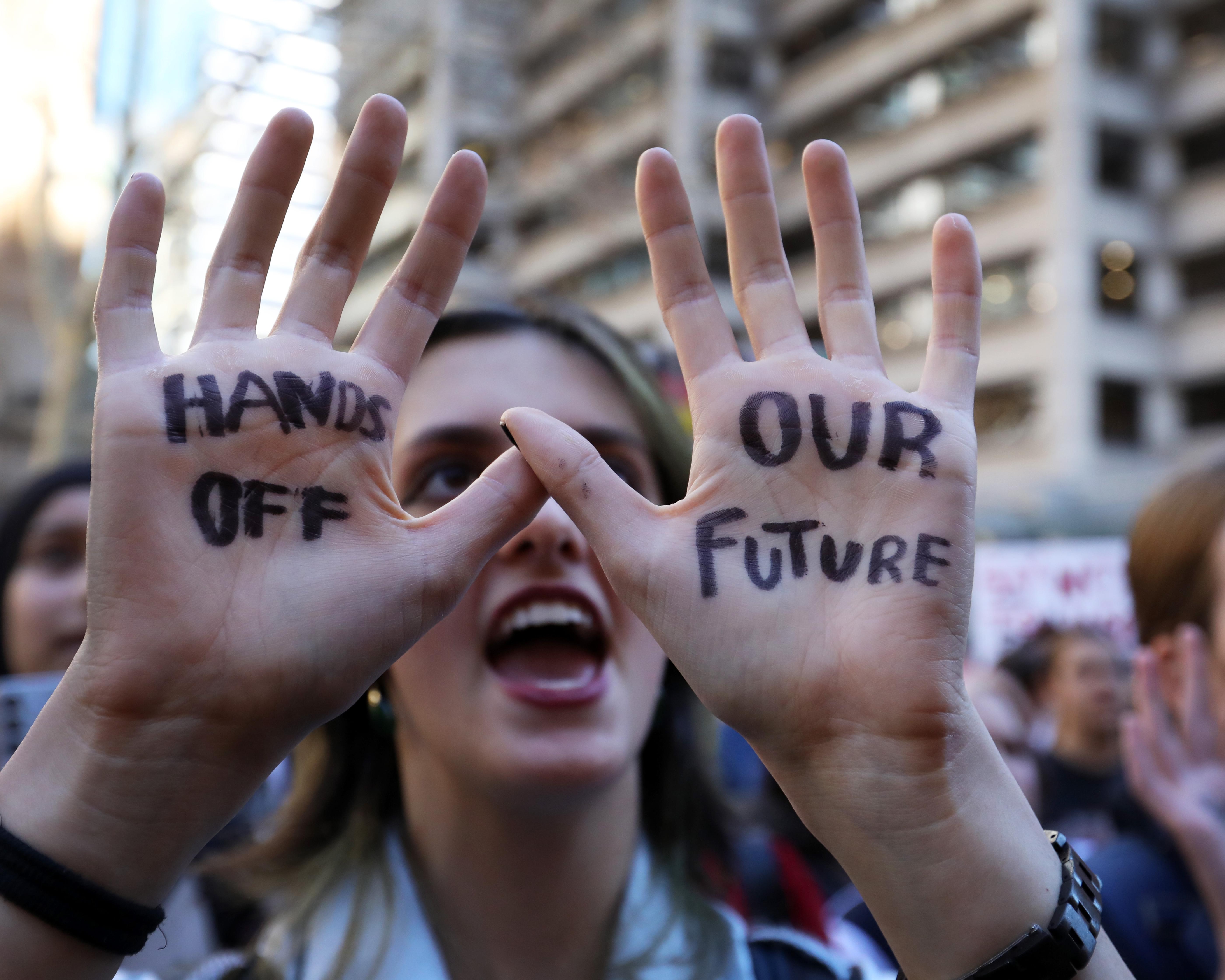 Protester raises hands marked “HANDS OFF OUR FUTURE” in front of a blurred crowd.