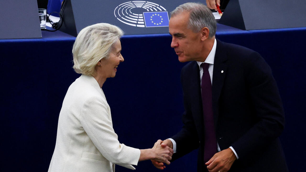 Two officials shake hands inside a European Parliament chamber, with EU symbols visible behind them.